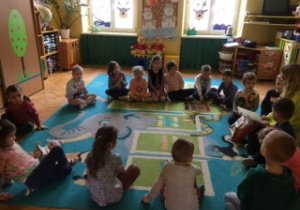 Dzieci siedzą na dywanie i słuchają czytanego tekstu.