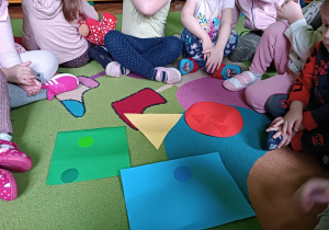 Dzieci siedzą na dywanie i dopasowują figury według koloru.