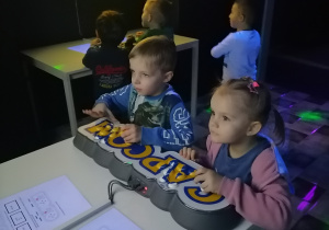 Dzieci stoją przy konsoli i patrzą na monitor.