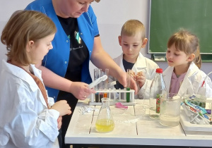 Dzieci stoją przy stole wraz z Panią Ewą wykonują eksperyment z kolorami i octem.