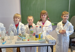 Dzieci stoją przy stole ubrane w białe fartuchy, aby wykonać eksperyment.