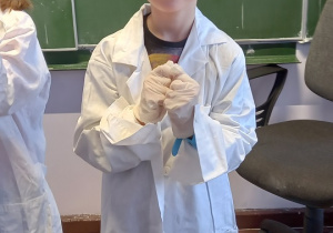 Chłopiec w białym fartuchu i gumowych rękawiczkach uśmiecha się do zdjęcia.