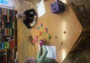 Dzieci siedzą przy stole z założonymi rękawiczkami i budują z klocków.