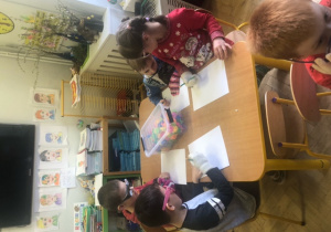 Dzieci siedzą przy stole z założonymi rękawiczkami i okularami. Rysują na kartkach.