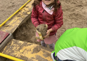 Dziewczynka stoi w piaskownicy i trzyma wiaderko.