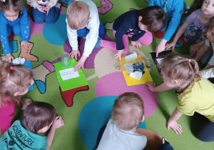 Dzieci siedzą na dywanie i dopasowują odpady do danego pojemnika.