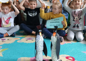 Dzieci siedzą i pozują do zdjęcia z wykonanymi pracami plastycznymi "Pingwin".