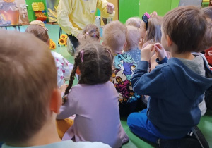Dzieci siedzą na dywanie i słuchają Pana w stroju ochronnym pszczelarskim.