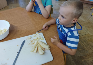 Chłopiec siedzi przy stoliku i kroi gruszkę na desce do krojenia.