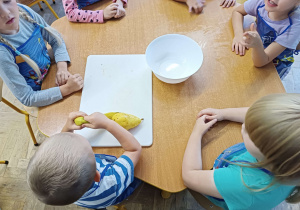 Zdjęcie dzieci przy stoliku. Na stoliku znajduje się miska i deska do krojenia z owocami.