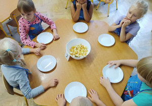 Zdjęcie dzieci przy stoliku. Na stoliku znajdują się talerzyki i miska z sałatką owocową.