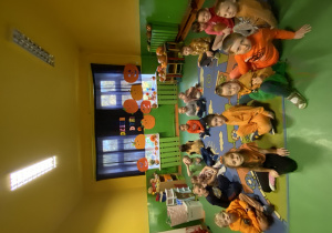Dzieci ubrane są w stroje koloru pomarańczowego i siedzą na dywanie.