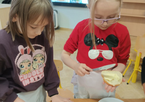 Dziewczynki stoją przy stole i smarują kromki chleba masłem.