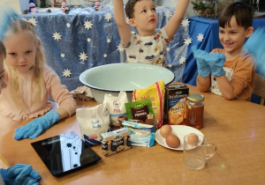 Dzieci siedzą przy stoliku z produkatami potrzebnymi do przygotowania pierniczków.