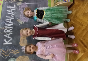 Trzy dziewczynki przebrane za księżniczki stoją pod napisem karnawał.