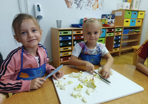 Uśmiechnięte dziewczynki siedzą przy stole i kroją jabłka na desce do krojenia.