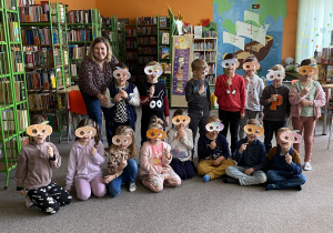 Dzieci siedzą i stoją w bibliotece. Prezentują swoje maski.