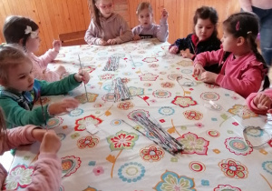 Dzieci siedzą przy stole i trzymają papierowe wałeczki.